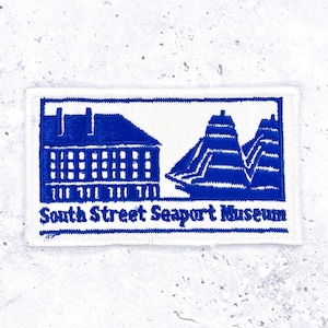 May include: A rectangular white patch with a blue embroidered design. The design features a building, a ship, and the text "South Street Seaport Museum" in blue.