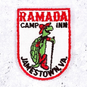 Peut inclure: Un écusson brodé blanc à bordure rouge représentant une tortue verte portant un chapeau et des bottes rouges, tenant un bâton de marche. Le texte indique RAMADA CAMP INN JAMESTOWN, VA en lettres noires et rouges sur fond blanc.