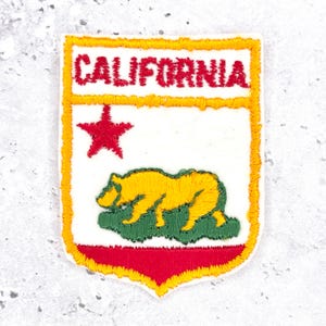 Może przedstawiać: Naszywka w kształcie tarczy z wyhaftowanym czerwonym napisem CALIFORNIA u góry. Poniżej znajduje się czerwona gwiazda po lewej stronie oraz żółty niedźwiedź kroczący po zielonej trawie. Całość wykończona jest grubą, żółtą nicią na krawędziach.