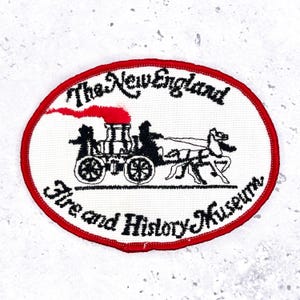 Op de afbeelding: Ovale patch met een rode rand en een witte kern. De patch toont een zwarte illustratie van een door paarden getrokken brandweerwagen met de tekst "The New England Fire and History Museum" in zwart schrift.