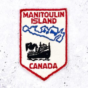 May include: An embroidered patch with a white background and a red border. The patch features the text "MANITOULIN ISLAND CANADA", a blue island outline, and a black ship.