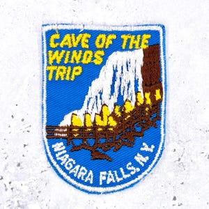 May include: A shield-shaped embroidered patch with a blue background featuring a white waterfall and brown wooden walkways. The yellow text reads CAVE OF THE WINDS TRIP and the white text at the bottom reads NIAGARA FALLS N.Y. with a white border.