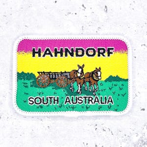 May include: Rectangular patch with a white border featuring the text "HAHNDORF" above "SOUTH AUSTRALIA." The patch has a gradient background of pink, yellow, and green. A brown horse-drawn cart is in the center.