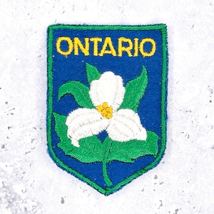 May include: Embroidered patch featuring the word "ONTARIO" in yellow above a white trillium flower with a yellow center and green leaves. The design is set against a blue background with a green border.