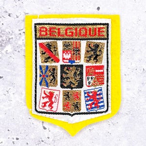 Può includere: Una toppa di feltro giallo con un disegno ricamato bianco con la scritta "BELGIQUE" in rosso e oro. Il disegno comprende più stemmi.