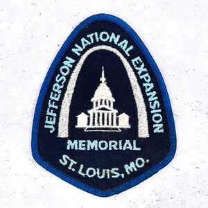 May include: A navy blue patch with a light blue border and a white and light blue design. The patch features the text "JEFFERSON NATIONAL EXPANSION MEMORIAL ST. LOUIS, MO." and an illustration of a building.