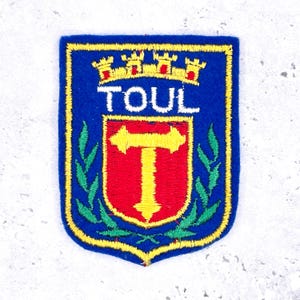 May include: A blue embroidered patch with a yellow border. The patch features the word "TOUL" above a red shield with a yellow "T" and a crown. Green leaves are on either side.