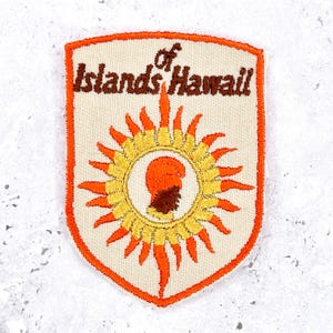 May include: A shield-shaped embroidered patch with an off-white background and a bright orange border. The text Islands of Hawaii is stitched in brown. A central yellow and orange sunburst design surrounds a brown silhouette of a human head.