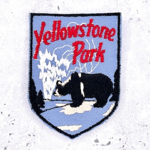 Może przedstawiać: Haftowana naszywka z czarnym niedźwiedziem, gejzerem i drzewami na jasnoniebieskim tle. Napis "Yellowstone Park" jest czerwony. Naszywka ma kształt tarczy z czarną obwódką.