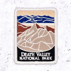 Può includere: Una toppa ricamata rettangolare che raffigura un paesaggio del Death Valley National Park. Il design include un terreno desertico color marrone chiaro, montagne marroni e colline blu scuro sotto un cielo color sabbia. Il testo DEATH VALLEY NATIONAL PARK è bianco.