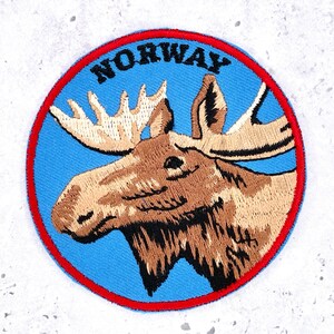 May include: A circular embroidered patch featuring a brown moose head against a light blue background. The word NORWAY is stitched in black capital letters at the top. The patch is bordered by a thin red stitched edge.