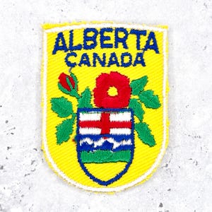 Könnte beinhalten: Ein gelber gestickter Aufnäher mit dem Text ALBERTA CANADA in Blau. Das Design zeigt eine rote Wildrose mit grünen Blättern und ein Wappen mit einem roten Kreuz, blauen Bergen und grünen Hügeln, umrandet von einem weißen Rand.
