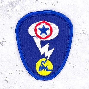 May include: A shield-shaped embroidered patch with a blue background and a dark blue stitched border. The design features a white oval with a red circular outline containing a blue star, a white lightning bolt, and a yellow circle with blue letters.