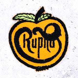 Op de afbeelding: Geborduurde patch in de vorm van een oranje appel, met zwarte contouren en het woord "Ruphus" in zwarte letters. De appel heeft groene bladeren en een zwarte steel. Geschikt voor kleding of accessoires.