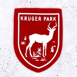 Può includere: Una toppa in feltro rosso a forma di scudo con un disegno ricamato in bianco di un'antilope nell'erba. Il testo KRUGER PARK è ricamato in bianco nella parte superiore della toppa, che è rifinita con un bordo bianco.