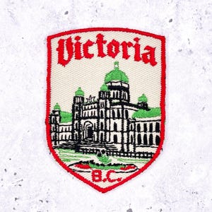 Könnte beinhalten: Gestickter Aufnäher mit dem Wort "Victoria" in roter Schrift über einer detaillierten Illustration eines Gebäudes mit grünen Kuppeln und Akzenten. Der Aufnäher hat einen roten Rand und die Buchstaben "B.C." unten.
