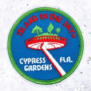 May include: A circular patch with a blue background and green border. The patch features the text "ISLAND IN THE SKY" in red, and "CYPRESS GARDENS FLA." in white. A red and white bridge design is in the center.