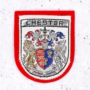Op de afbeelding: Geborduurde patch met een rode rand en een gedetailleerd wapenontwerp. Het wapen heeft een schild met rode en blauwe secties, een zwaard en het woord "CHESTER" bovenaan. De patch bevat de Latijnse zin "ANTIQUI COLANT ANTIQUUM".