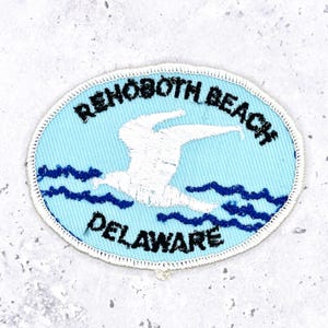 May include: Oval patch with a light blue background featuring a white seagull soaring above dark blue waves. The patch is embroidered with the words "REHOBOTH BEACH DELAWARE" in black.