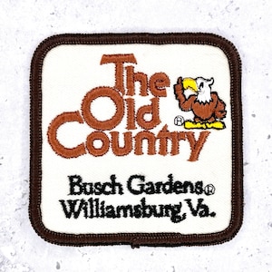 May include: A square embroidered patch with a white background and a dark brown border. It features the brown text The Old Country, a graphic of a brown and white eagle, and the black text Busch Gardens Williamsburg, Va. below the eagle.