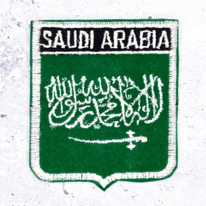 May include: A shield-shaped embroidered patch featuring a green background with a white border. The text SAUDI ARABIA is stitched in white on a black horizontal band at the top. Below, white Arabic calligraphy and a sword are embroidered.