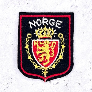 May include: A black embroidered patch featuring a red shield with a yellow lion holding an axe, topped by a yellow crown. The word NORGE is stitched in white above the shield, and the entire design is bordered by a red line on a black background.
