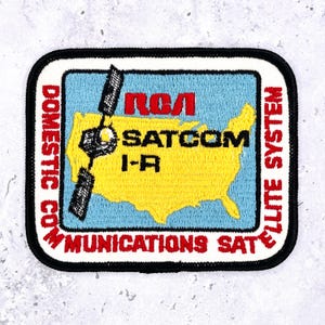 May include: Embroidered patch featuring a satellite design and the text "RCA SATCOM I-R." The patch has a blue background with a yellow outline of the United States. The border includes the words "DOMESTIC COMMUNICATIONS SATELLITE SYSTEM."