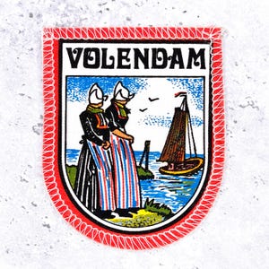 Op de afbeelding: Een schildvormige geborduurde patch met een rode gestikte rand. Het ontwerp toont de tekst VOLENDAM in zwarte letters boven een illustratie van twee figuren in traditionele Nederlandse klederdracht bij een blauwe zee met een bruine zeilboot.