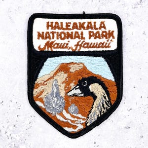 May include: Embroidered patch featuring the text "HALEAKALA NATIONAL PARK Maui, Hawaii." The patch has a black border and depicts a mountain scene with a goose. The colors include brown, blue, white, and black.