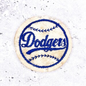 Può includere: Una toppa ricamata circolare con uno sfondo color crema, un motivo a cuciture da baseball cucito in blu e la parola Dodgers scritta in corsivo blu al centro, che imita il look classico del logo di una squadra di baseball professionistica.