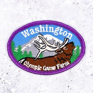 May include: An oval embroidered patch with a purple border featuring a brown elk with large white antlers in front of a snow-capped mountain and green pine trees. The text Washington is at the top and Olympic Game Farm is at the bottom.