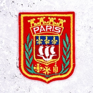 Può includere: Toppa ricamata con uno scudo rosso con la scritta "PARIS" in bianco. La toppa include un emblema blu e oro, una nave bianca e foglie di alloro verdi. Il bordo è giallo.
