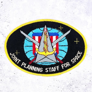 May include: Embroidered patch with a black oval border and yellow stitching. The patch features a globe with a red, white, and blue design, crossed swords, and an anchor. Text reads "JOINT PLANNING STAFF FOR SPACE".