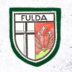 May include: A patch with a green border and a white, red, and black design. The word "FULDA" is at the top. The design includes a cross and three white flowers.