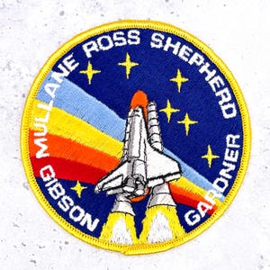 May include: A circular embroidered patch featuring a space shuttle launching against a dark blue background with yellow stars. A rainbow-colored trail extends behind the shuttle. The text MULLANE ROSS SHEPHERD GIBSON GARDNER is stitched in white.