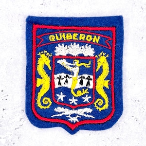 May include: A blue felt patch featuring a red border and the text QUIBERON in yellow. The design includes two yellow seahorses flanking a central shield with a yellow mermaid figure, white stars, and white stylized figures on a blue background.