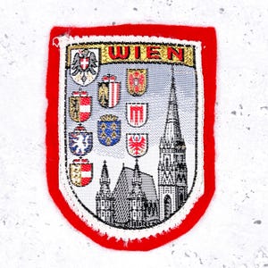 May include: A rectangular patch with a red border and the word "WIEN" in gold letters. The patch features a depiction of a church and several crests, with a white background.