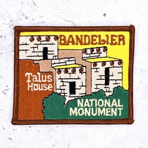 May include: A rectangular embroidered patch featuring a yellow background with a brown border. It depicts the Bandelier Talus House National Monument with white stone structures, dark brown accents, and green foliage. The text reads Bandelier, Talus House, and National Monument.