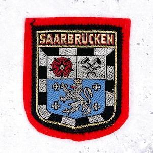May include: A rectangular patch with a red border and a detailed emblem. The emblem features the word "SAARBRÜCKEN" in gold lettering, a checkered pattern, a red rose, crossed tools, a blue shield with a lion, and decorative crosses.