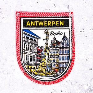 Pode incluir: Um patch retangular com uma borda vermelha e a palavra "ANTWERPEN" em letras amarelas. O patch apresenta uma ilustração detalhada de edifícios e uma estátua, com a palavra "Brabo" acima da estátua.