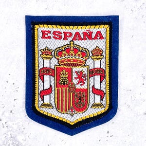 May include: A rectangular embroidered patch with a blue felt border featuring the coat of arms of Spain. The design includes a central shield, a crown, and two pillars with red ribbons. The word ESPAÑA is stitched in red at the top.
