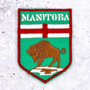 May include: Embroidered patch featuring the word "MANITOBA" above a red cross on a green background. A brown bison stands on a white and green base. The patch has a red border.