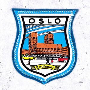 May include: A shield-shaped embroidered patch featuring a graphic illustration of the Oslo City Hall building. The design includes the text OSLO at the top and Rådhuset on a yellow banner at the bottom, with blue, orange, and black colors.