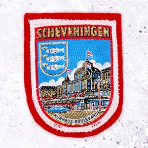 Op de afbeelding: Een rechthoekige patch met een rode rand en witte stiksels. De patch toont het woord "Scheveningen" in gouden letters boven een blauw schild met drie vissen. Daaronder een gedetailleerde illustratie van gebouwen en de tekst "KURHAUS-BOULEVARD".