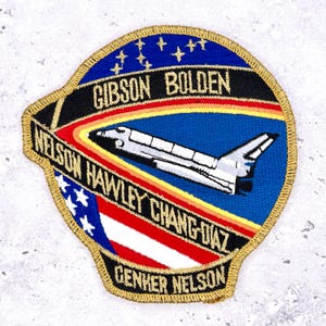 May include: Embroidered patch featuring a space shuttle design. The patch has a blue background with a rainbow arc and the names GIBSON BOLDEN, NELSON HAWLEY CHANG-DIAZ, and GENKER NELSON. The patch is edged with gold.