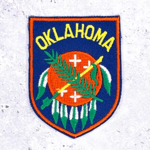 May include: Embroidered patch featuring the word "OKLAHOMA" in yellow arched over a shield-shaped design. The design includes a central orange circle with white crosses, a green sprig, and crossed yellow staffs. The patch has a blue background and orange border.