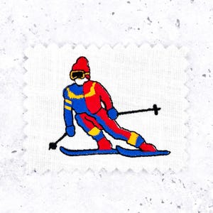 May include: Embroidered patch depicting a skier in a downhill pose. The skier wears a red and blue ski suit with yellow details, black ski poles, and blue skis. The patch is on a white, square fabric.