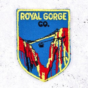 May include: Embroidered patch with a blue background and yellow border. The patch features the text "ROYAL GORGE CO." above a graphic of a mountain range with a cable car. Red and yellow accents highlight the mountain.