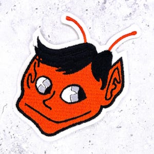 May include: An embroidered patch featuring a stylized orange devil head with black hair, two orange antennae, and large white eyes with black pupils. The design is outlined in black thread and set against a white fabric backing with a clean border.