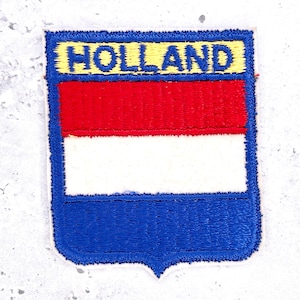 May include: A rectangular embroidered patch featuring a shield-like bottom edge. The design displays the word HOLLAND in blue letters on a yellow background above horizontal red, white, and blue stripes, all bordered by a thick blue stitched edge.